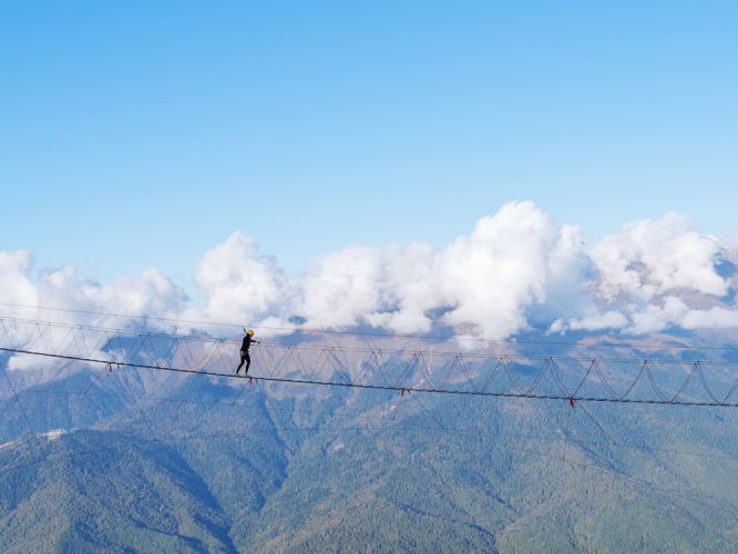 A girl in a helmet crosses a suspension bridge over a mountain gorge on clear day